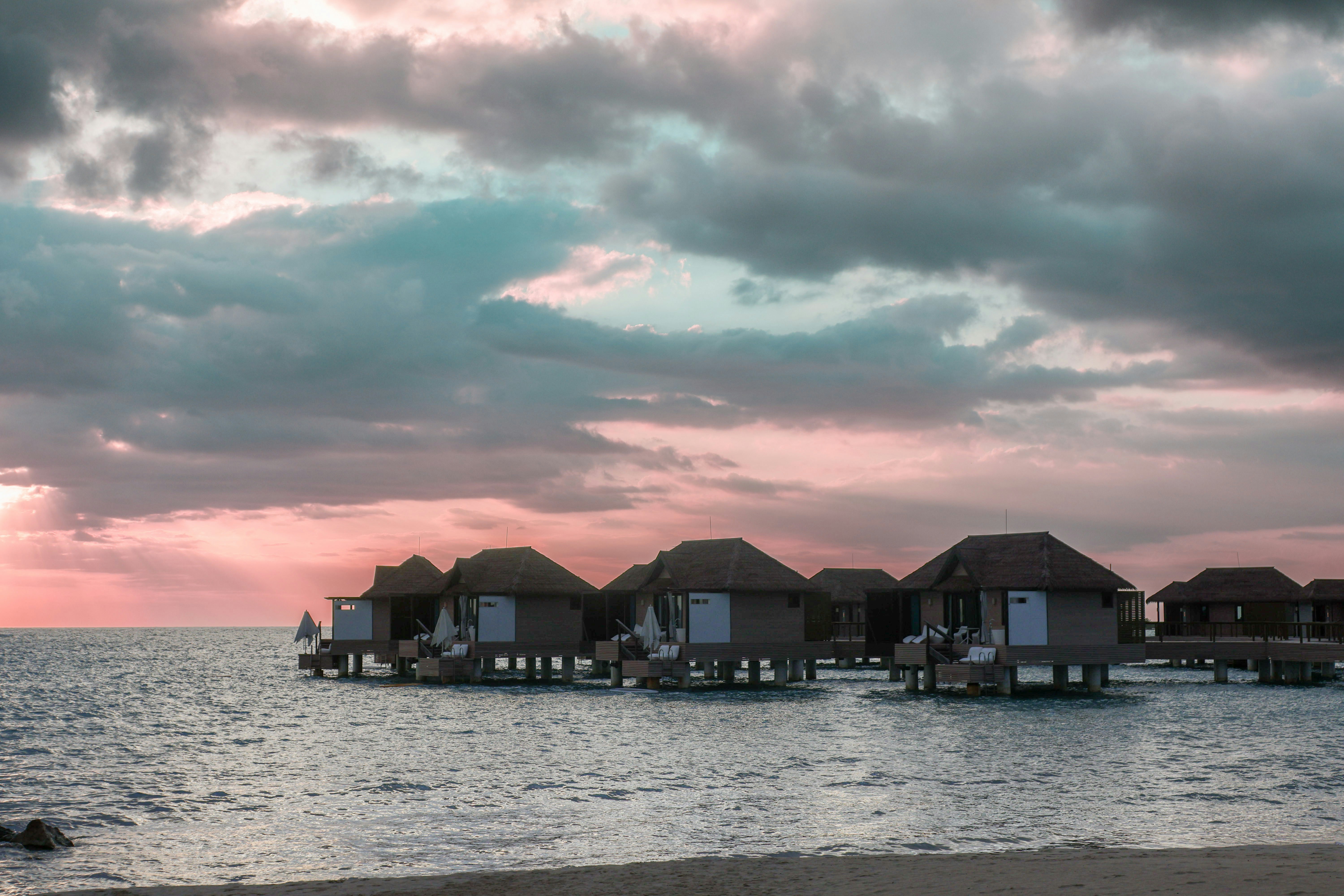 Sandals South Coast overwater bungalows on a wooden pier at sunset, with private decks and dramatic Caribbean sky — no volcanic peaks